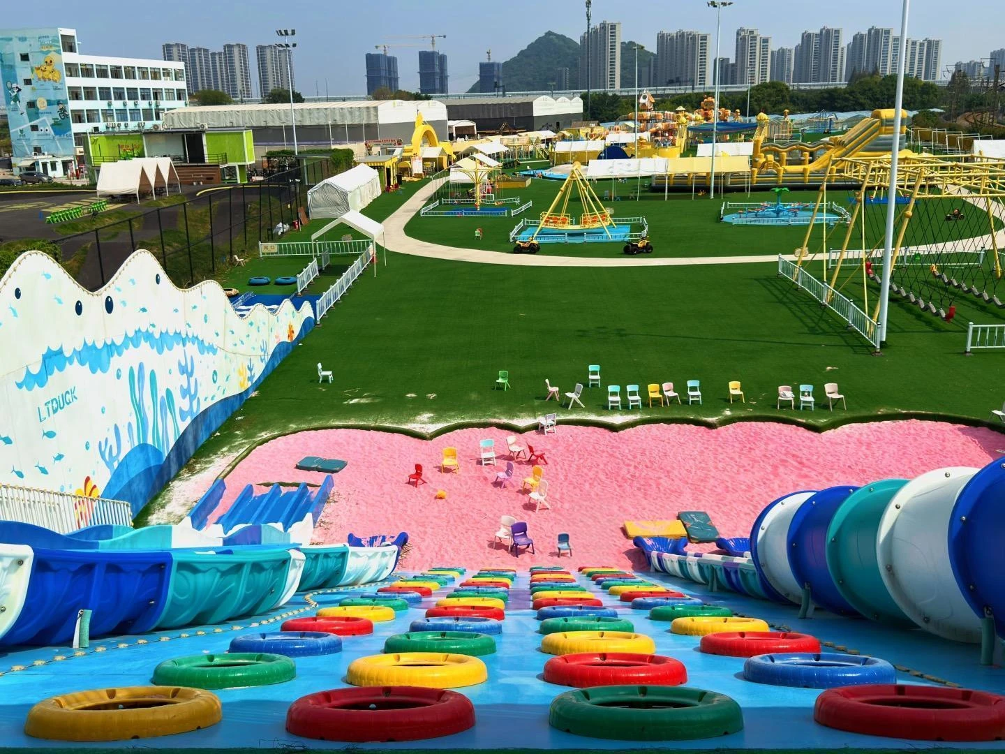 Elevated view of an outdoor leisure park with a foreground sliding area, circular ride tubes, swing structures and activity zones linked by pale paths.