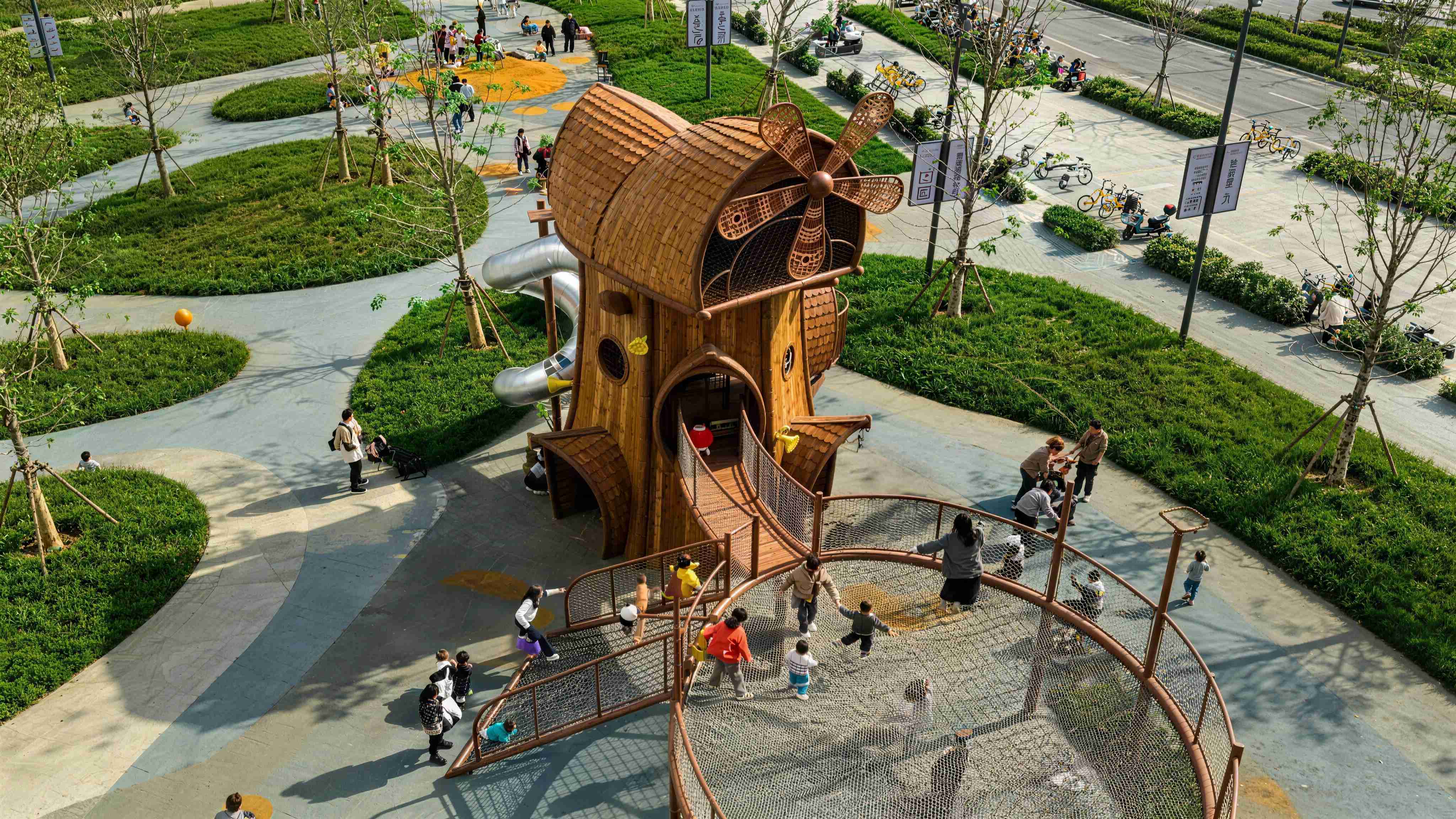 Durable custom commercial outdoor playground equipment featuring a large wooden windmill-themed playhouse, a stainless steel tube slide, and a heavy-duty suspended climbing net installed in a community park.